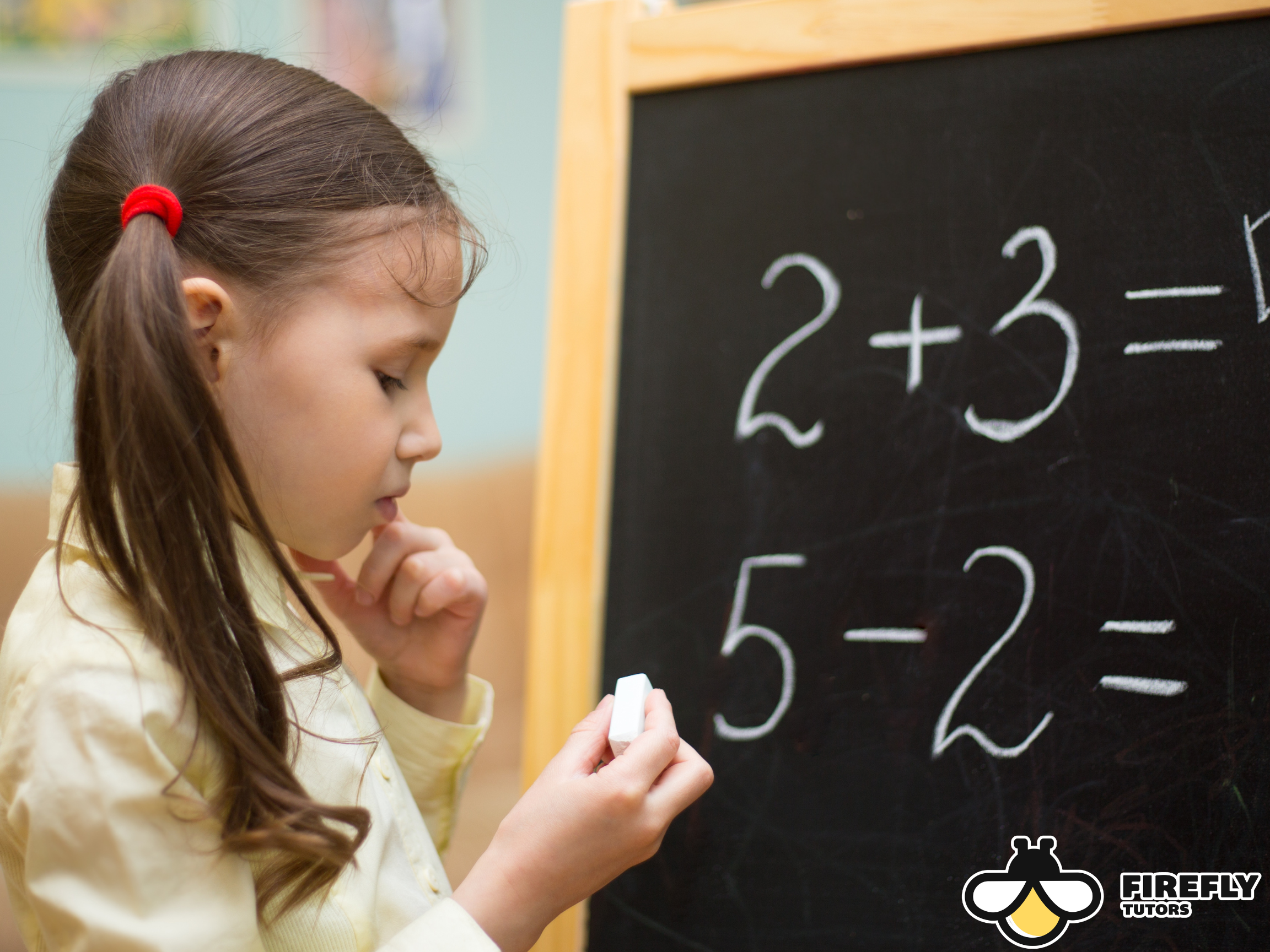 Young girl thinking while writing math problems on a chalkboard in a classroom setting. Math tutoring for kids