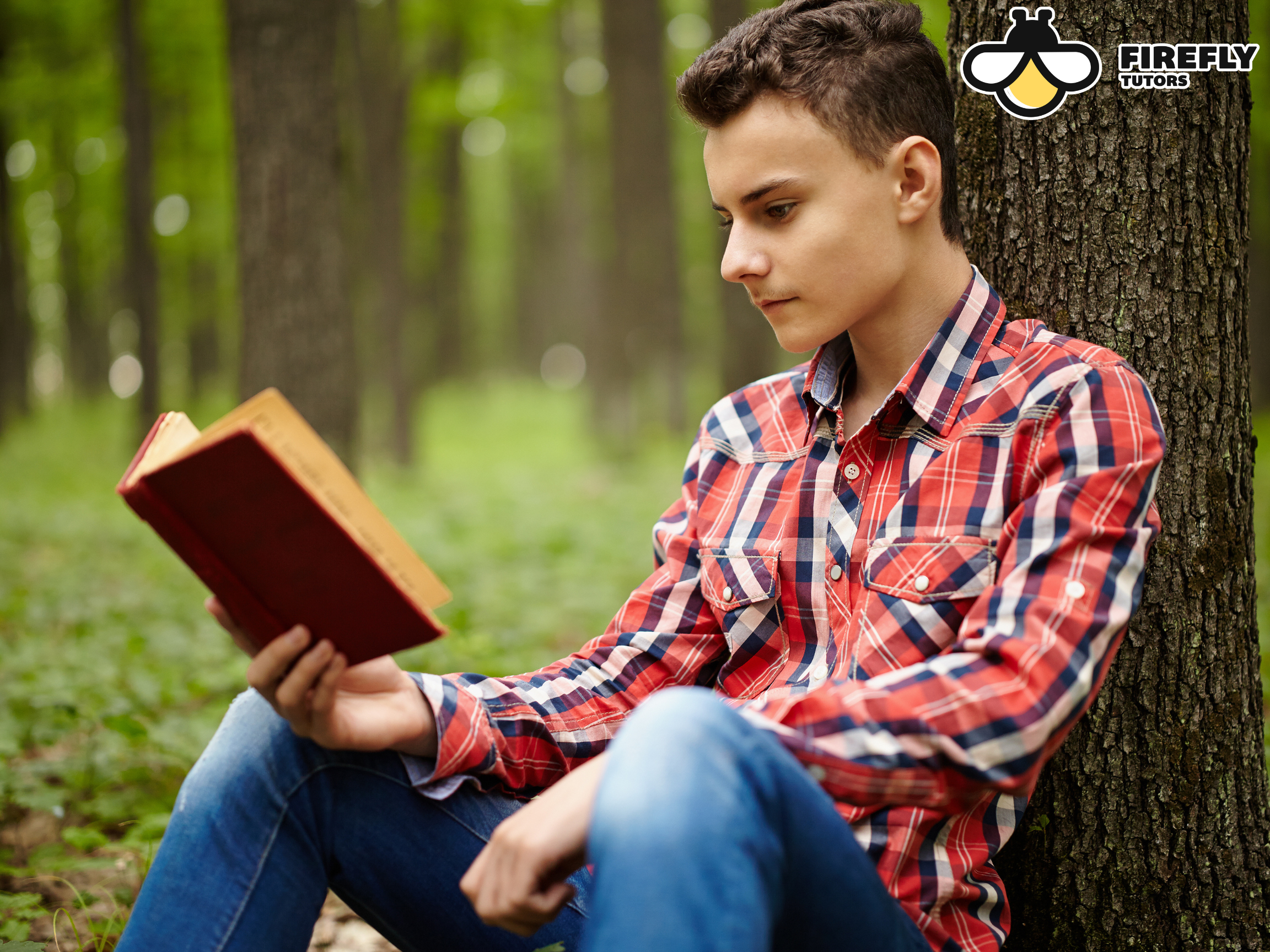 Teen boy leaning against a tree reading a book outdoors in summer