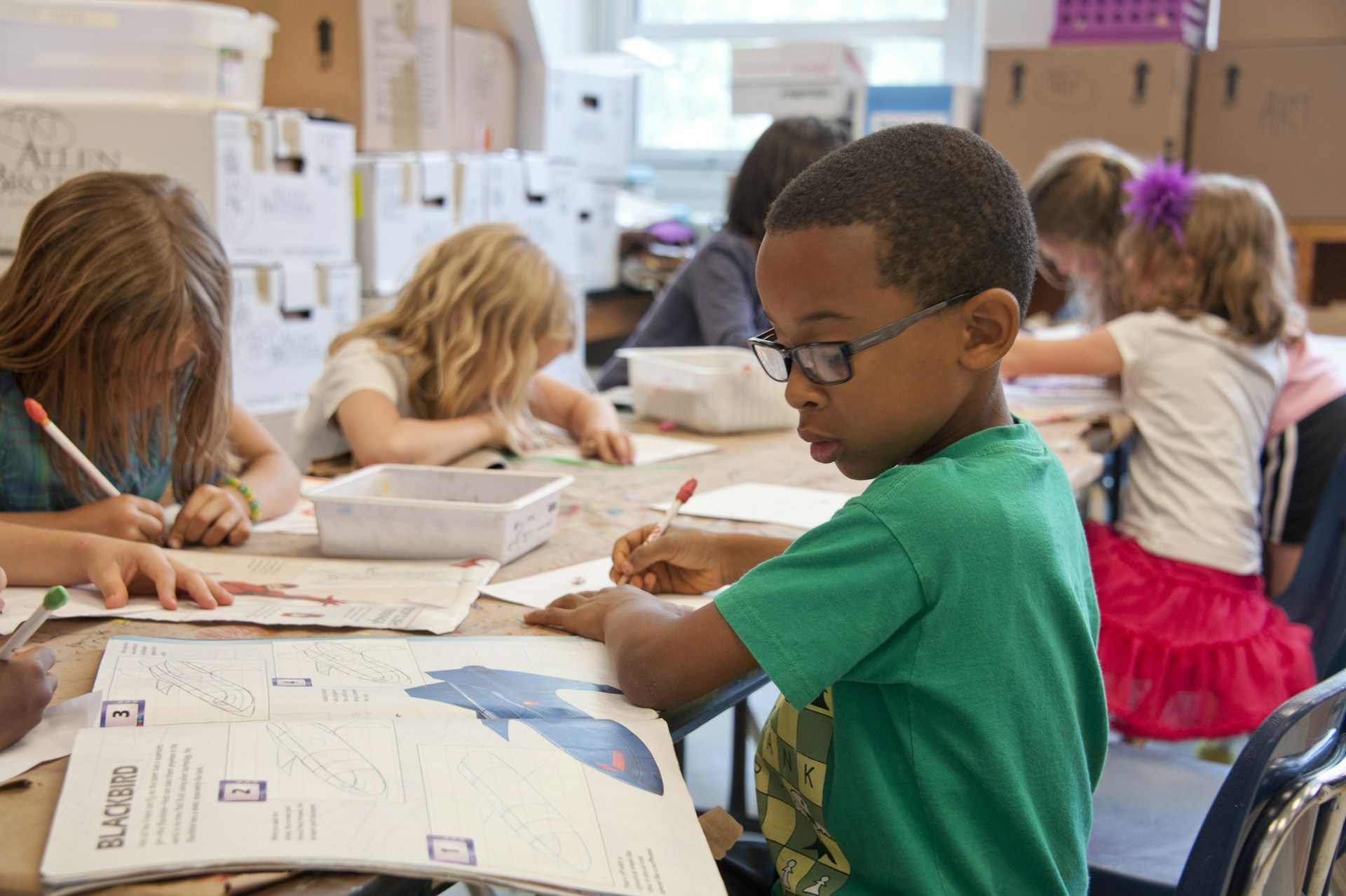Children in a classroom work quietly on art projects at a shared table, while a boy in glasses writes in the foreground.